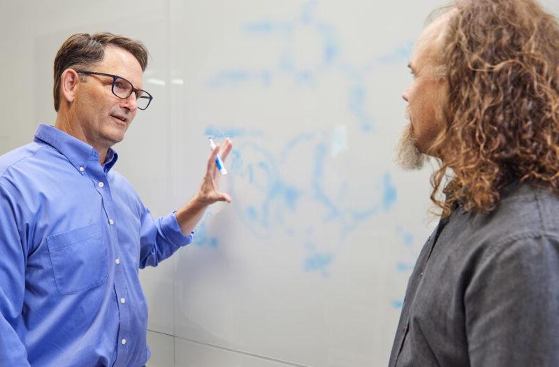 澳门正规博彩十大排行平台 scientists Brett Crawford and Dan Wendt talking by a whiteboard with a scientific formula